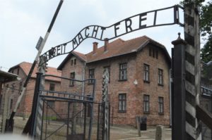 a gate with a sign in front of a brick building with Auschwitz concentration camp in the background