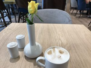 a white vase with a yellow flower in it next to a white mug with a foamy foam