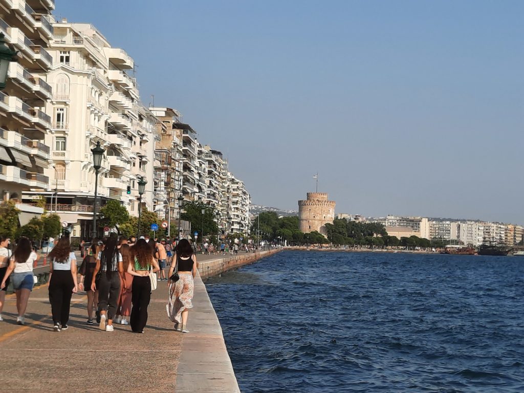 a group of people walking along a waterfront with Thessaloniki in the background