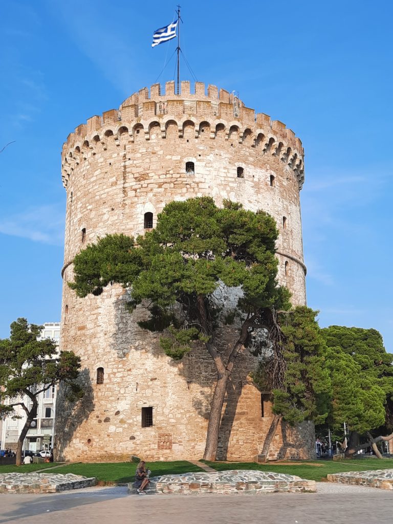 a stone tower with trees around it with White Tower of Thessaloniki in the background